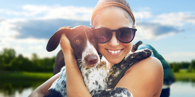 Young woman wearing sunglasses is smiling and holding her brown and white dog close to her face