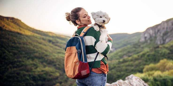 A woman hikes in the mountains holding a small white dog