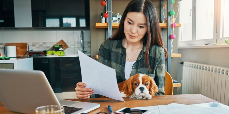 Woman sitting at a kitchen table looking at paperwork with a dog in her lap
